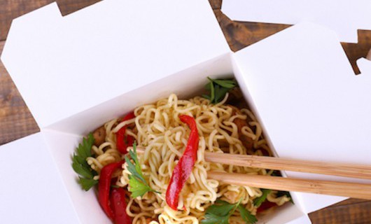 Two white cardboard takeaway boxes, containing fresh noodles and vegetables, are on a wooden table.