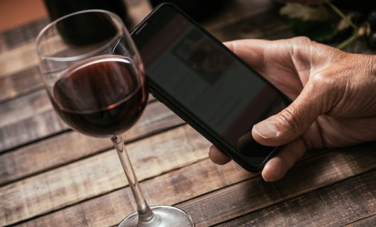 A hand holds a smart phone at a wooden table, with a small glass of red wine next to it.