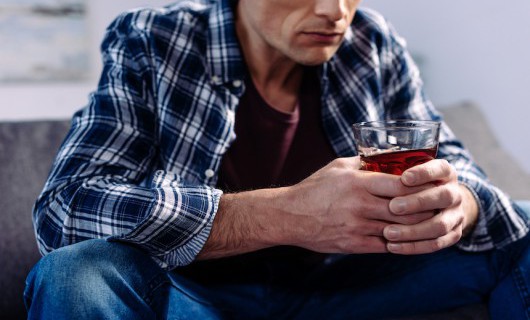 A man in a blue checked shirt sits against a grey step, holding a glass of alcohol.