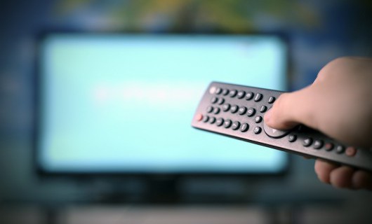 A hand is holding a television remote control, pointing it at an out of focus television screen.
