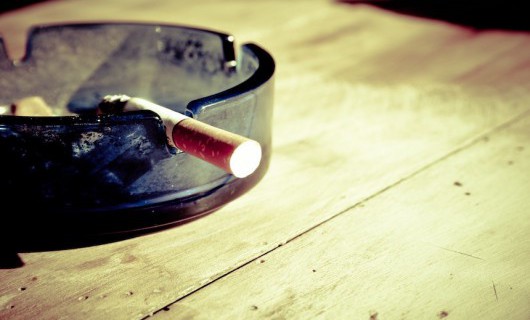 An ashtray is on a wooden table, with a half smoked cigarette hanging out of the ashtray.