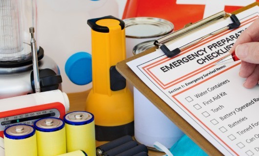 A person holding a clipboard with an emergency plan attached to it in front of various emergency supplies.