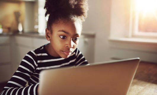 A young girl is looking at and using an open laptop on her kitchen table.