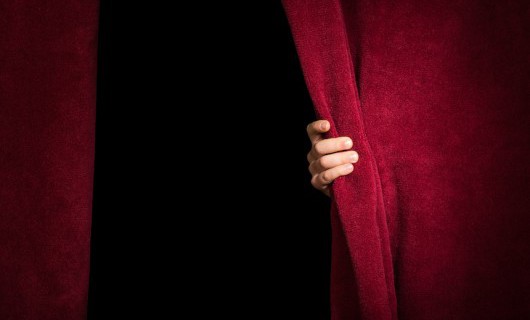 A pair of red velvet stage curtains, with a hand holding the right curtain back slightly.