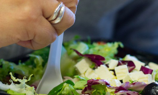 A hand is holding a plastic fork in a plastic container which is full of fresh salad.