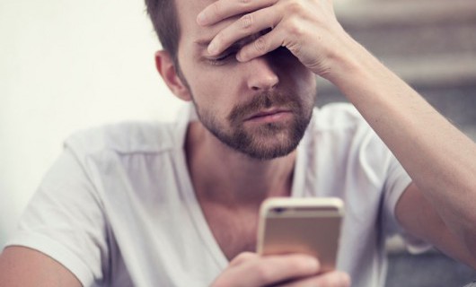 A man is looking at his smartphone whilst holding his head in his hand.