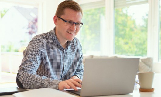 A man is smiling and looking engagingly towards his open laptop.