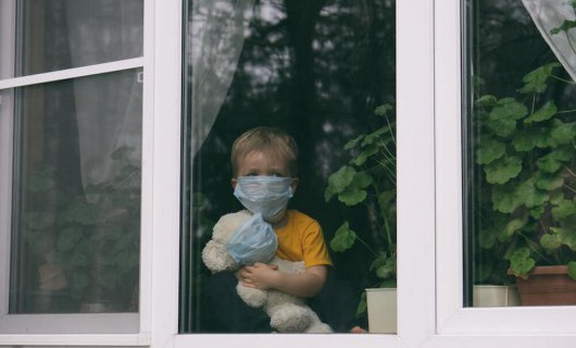 A young boy holds a teddy and stares out of a window. Both are wearing face masks.