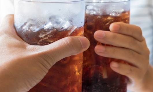 Two people clinking large glasses at a table, which are full of Coke, a straw and ice cubes.