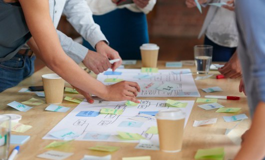 People are gathered around a table covered with sticky notes, diagrams and drinks.