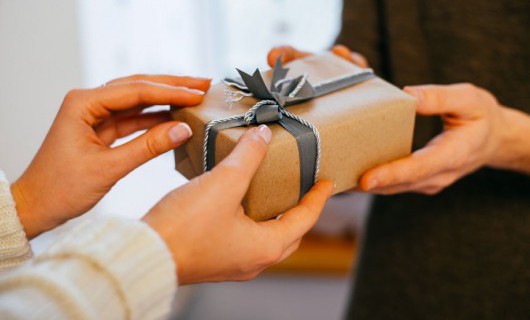 Two people are exchanging a gift, wrapped in brown paper with a black ribbon.