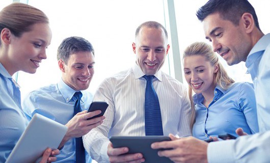 A group of professionals holding different devices smile down at an electronic tablet.