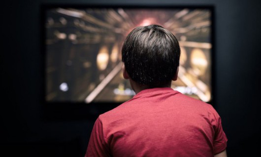 From behind, a young boy is playing a video game on his television in a dark room.