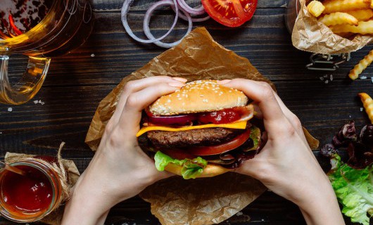 Two hands hold a large burger on a wooden table top, surrounded by a pint glass, sauces and chips.