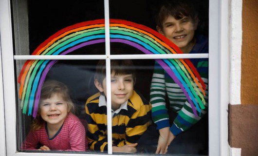 Three young children are smiling through a window painted with a rainbow.