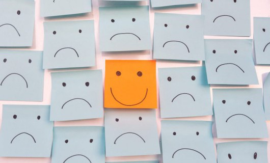 Numerous blue sticky notes with a sad face placed surround one orange note with a smile.