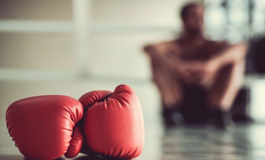 Behind a red pair of boxing gloves, someone sits on the floor with their knees pulled high.
