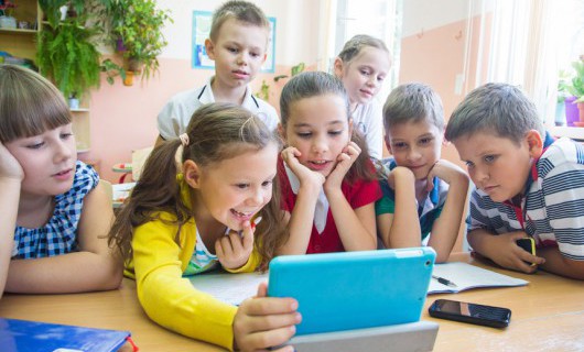 A group of young school children, some holding mobiles, playing with a tablet computer.