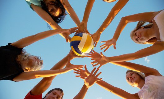 Six young girls are looking down at the camera and reaching towards a volleyball.