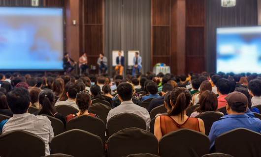 In a conference hall, the audience looks towards five people giving a presentation.