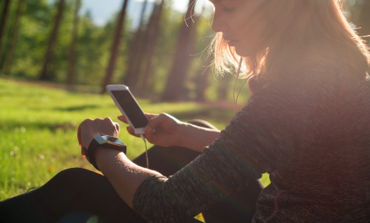 A woman sitting on the grass looks at her smartphone and smartwatch.