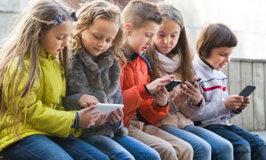 A group of children are sat on a bench looking at and playing on smartphones and tablets.