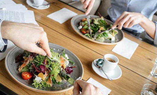 Two sets of hands eat colourful salads on a wooden table.