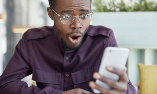 A man wearing glasses is staring at his smartphone with his mouth open in shock.