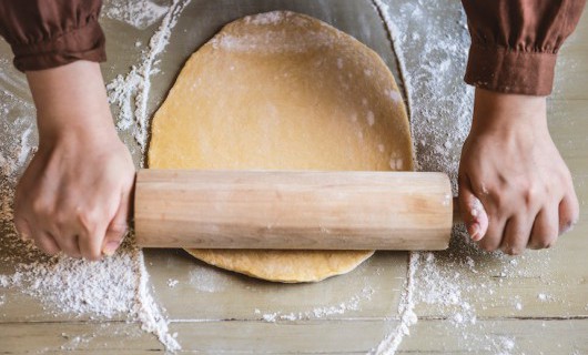 Seen from above, hands are using a rolling pin to roll out dough on a floured surface.