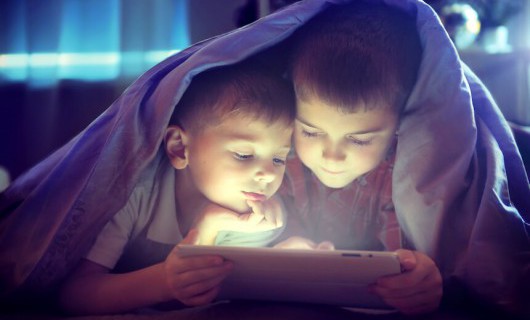 Two children play on a tablet computer underneath a blanket in a dimly lit bedroom.