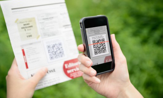 A pair of hands holding a mobile phone is scanning a QR code on a page held in front of the phone.