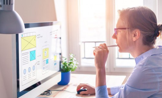 A woman chewing her pen looks at a computer displaying mock-ups for a phone app.