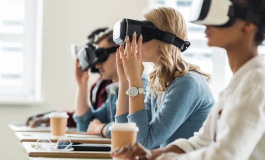 Four people are sat in a row, at separate desks and are wearing virtual reality headsets.