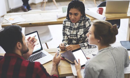 Three young people are in discussion, working with various devices and documents.