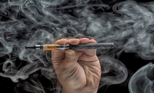 A hand holds a black e-cigarette horizontally, which is surrounded by smoke.