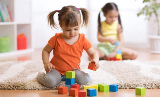 Two small girls sit on a rug at home, building towers with the colourful wooden blocks.