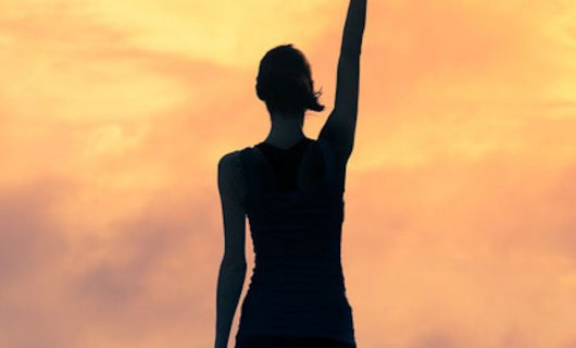 The silhouette of a woman against an orange sunset sky. She is in a power stance, punching the air.