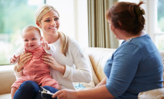 A healthcare worker sits on a sofa with a young woman who is holding a baby on her knee and smiling.
