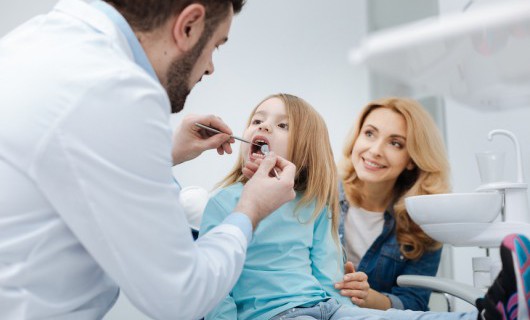 A dentist is checking a young girl's teeth, whose mother smiles behind them.