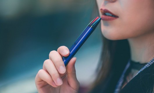 The bottom half of a young woman's face is shown as she holds an e-cigarette to her lips.