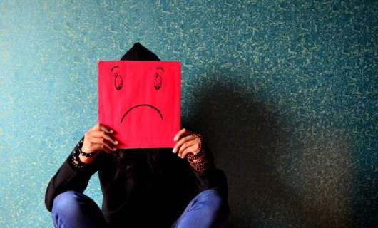 A teenager sits on the floor, covering their face with a sad face drawn on a red piece of card.