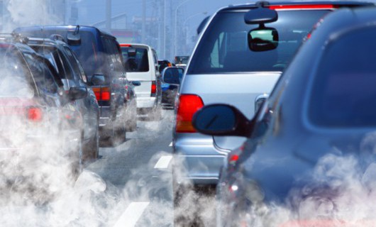 Two lines of vehicles are queuing in a traffic jam, with clouds of fumes coming from their exhausts.