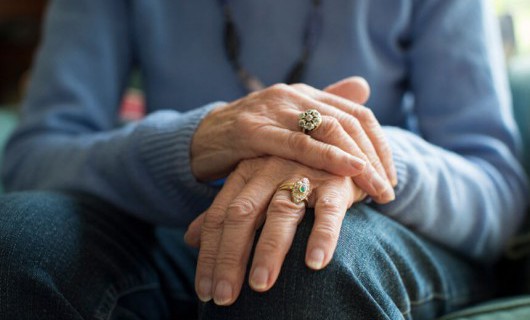 An elderly person's hand are on top of each other on their knee, one of which has many rings.