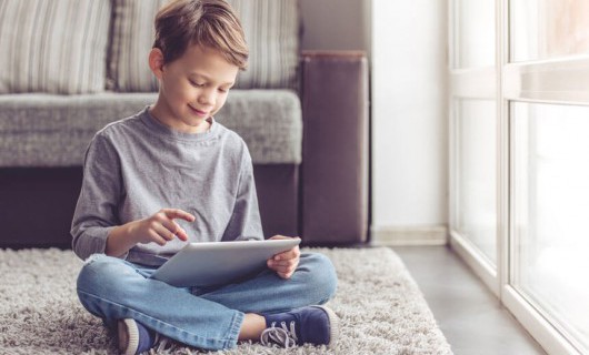 A young boy is sat on a rug at home smiling down at an iPad in his hands.