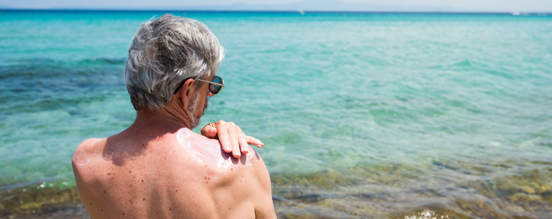 An older man wearing sunglasses applies suntan lotion to his upper back at a beach.