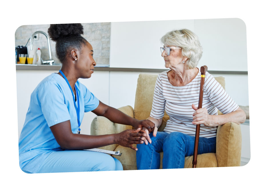 A female caregiver holding the hand of her elderly care-user who sits in an armchair as they speak.