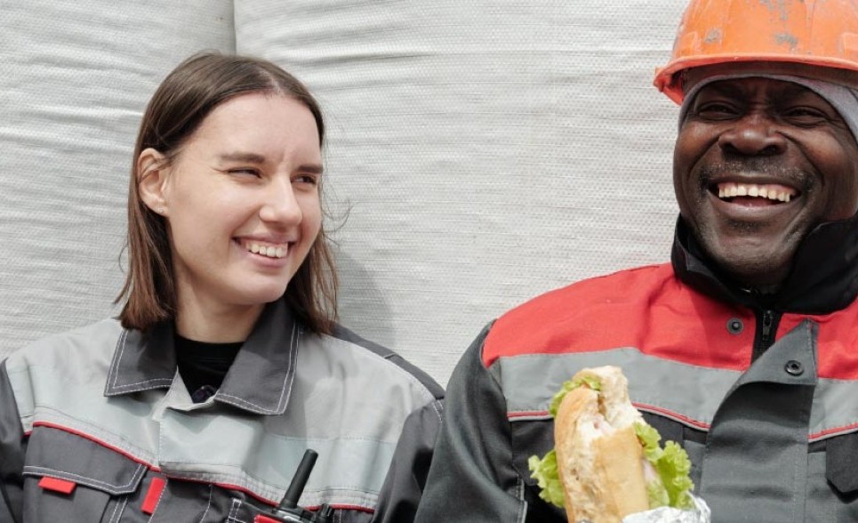Two construction workers sit eating lunch, they are laughing and smiling.