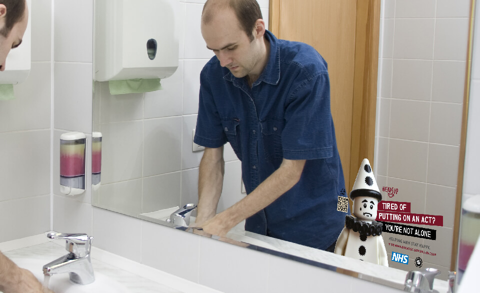 A man washing his hands in a public toilet which has a Heads-Up campaign decal on the mirror.