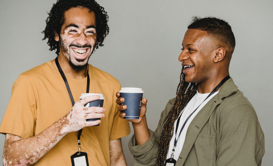 Two men laugh whilst holding cups of coffee, the man on the left lives with Vitiligo affecting parts of his face and right arm.