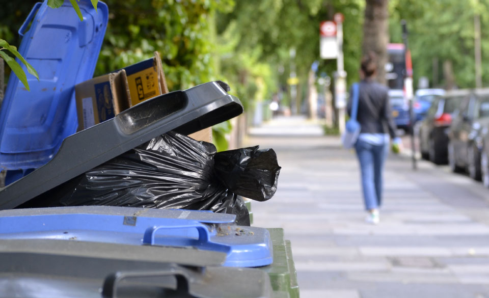 A close up of several wheelie bins over flowing with the amount of black bin bags inside them. A woman walks down the street in the distance beside a line of parked cars.